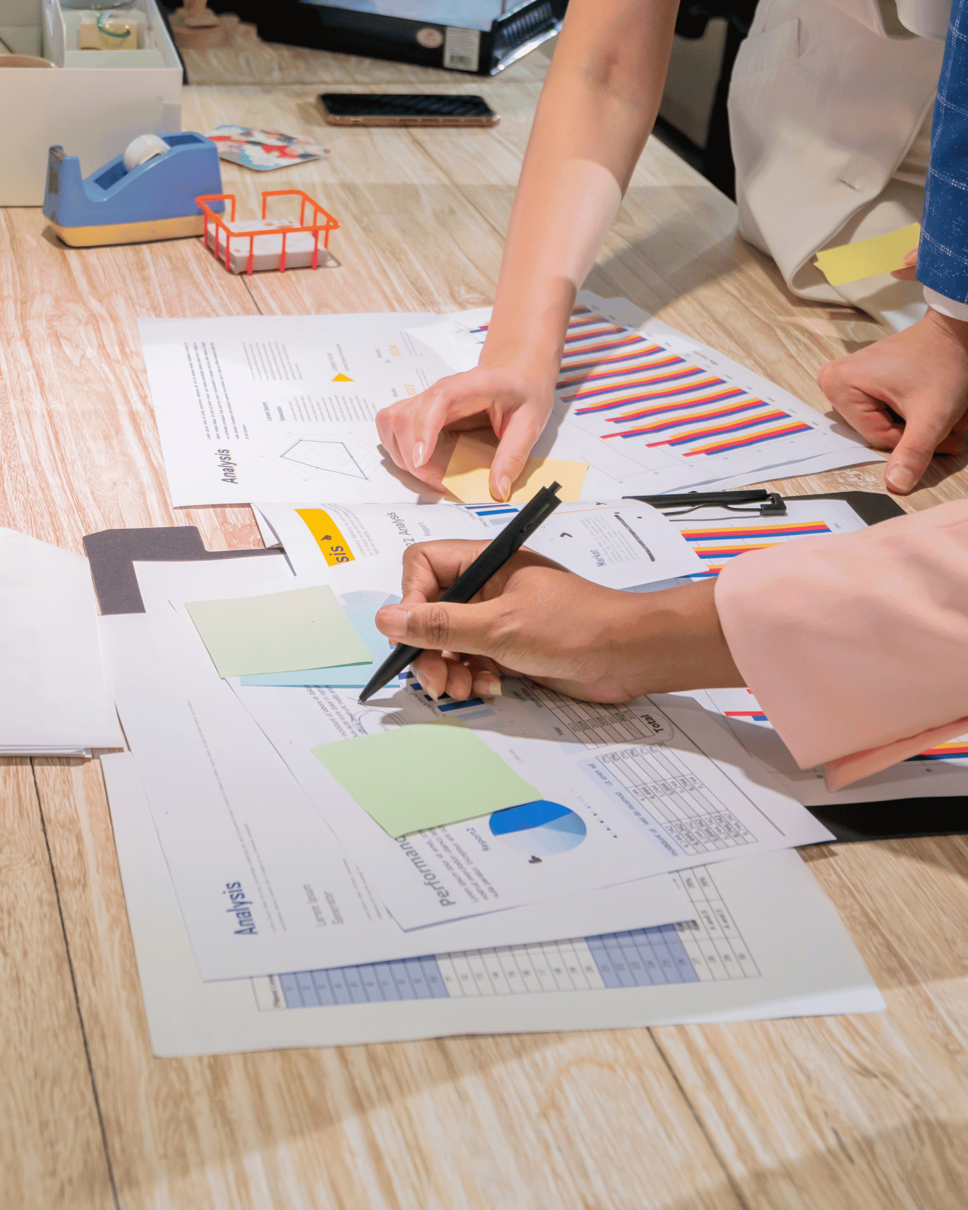 Golwg uwchben o fwrdd pren gyda dogfennau busnes, graffiau a nodiadau gludiog, wrth i dri o bobl gydweithio i ysgrifennu, pwyntio a dal nodyn.