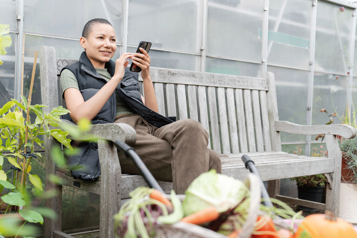 Menyw yn eistedd ar fainc yn defnyddio'i ffôn mewn rhandir.